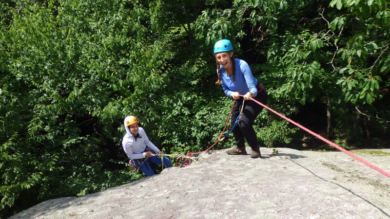 Two people are rappelling down a rock face. The person in the foreground is standing and holding the rope, while the other person is lower down, also holding the rope. Both are wearing helmets and dark-colored pants. The background is filled with green trees and foliage. The sky is not visible.